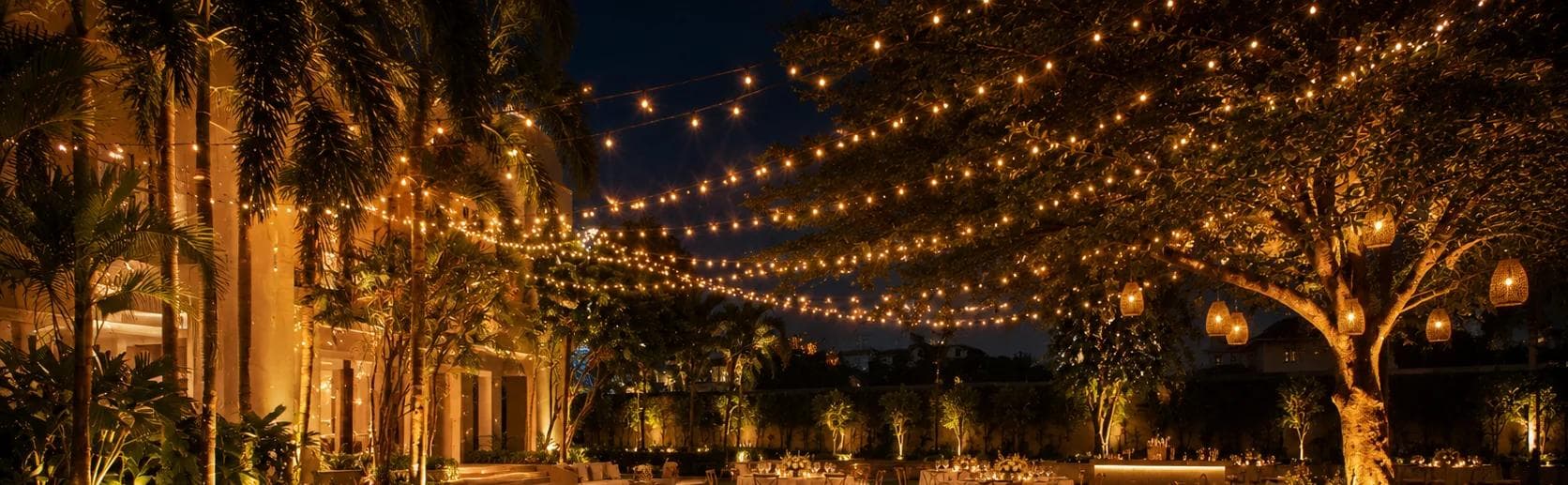 A garden reception at night, tables lit beneath trees strung with string lights and hanging lanterns.